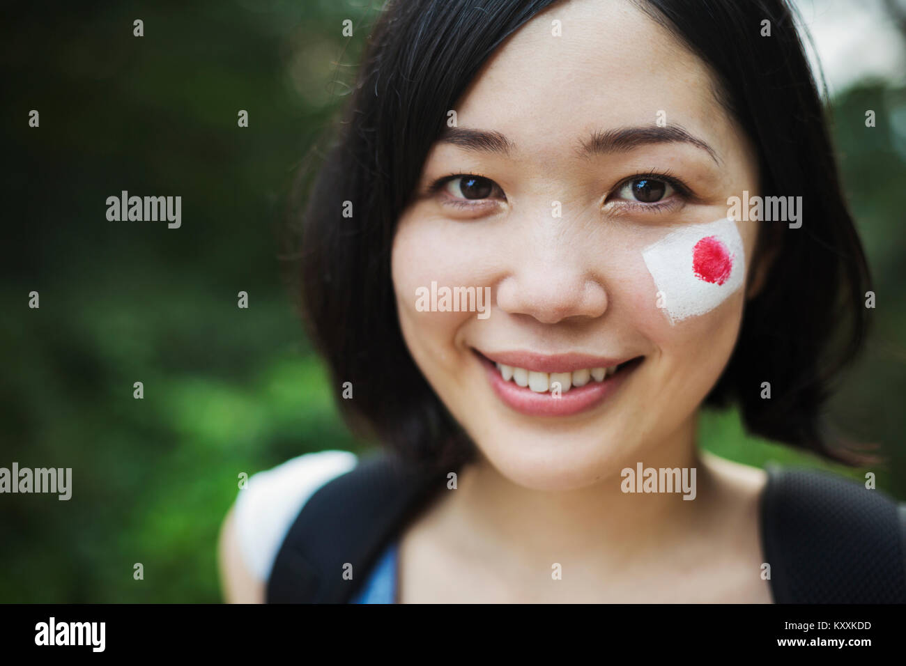 Ritratto di giovane donna con i capelli neri, bandiera giapponese dipinta sulla sua guancia, sorridente alla fotocamera. Foto Stock