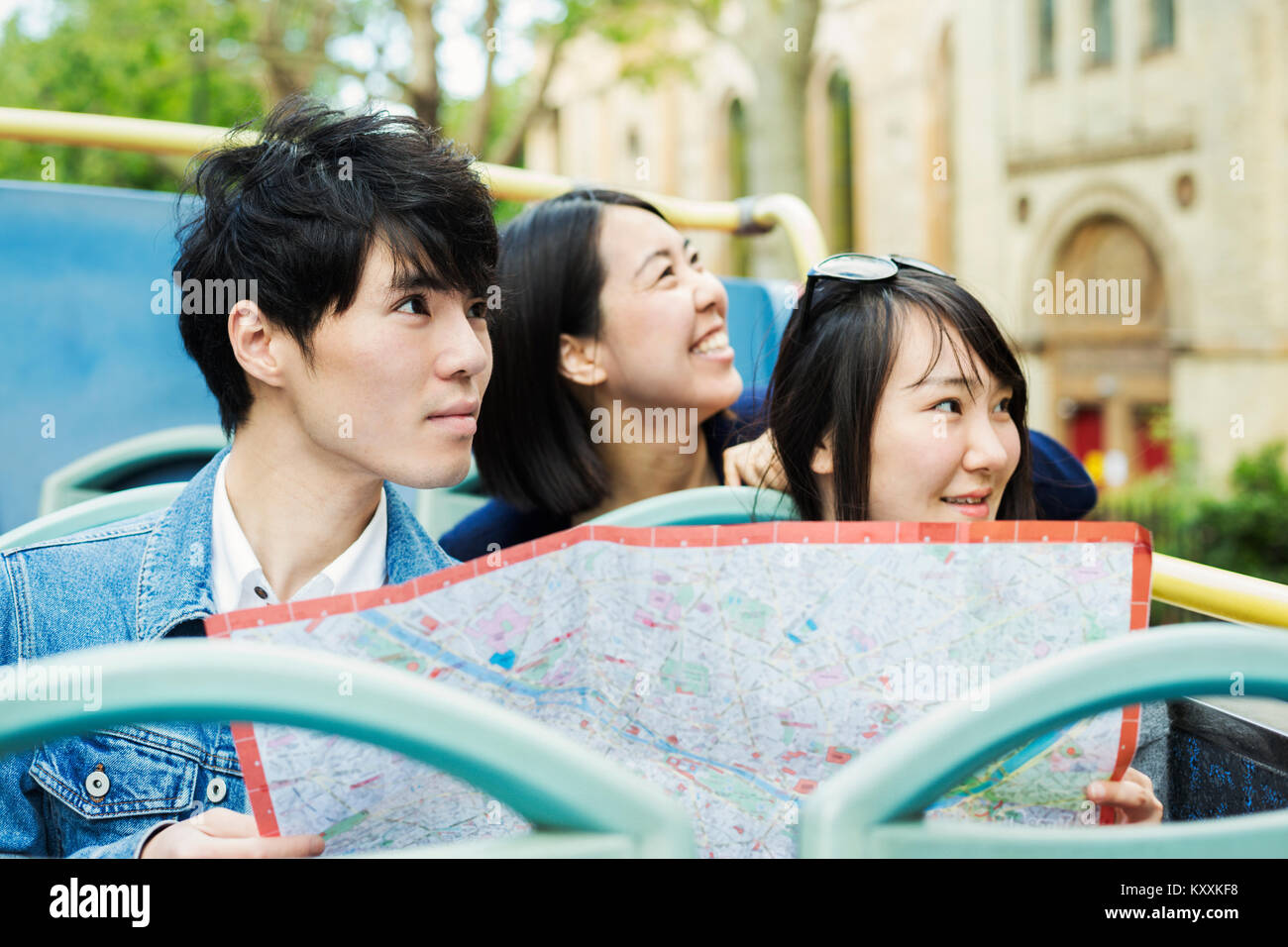 Uomo sorridente e due donne con capelli neri azienda mappa della città, seduto sulla cima di un open bus Double-Decker, guidando lungo la strada urbana. Foto Stock