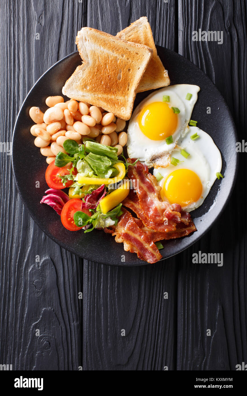 Deliziose uova fritte, pancetta, fagioli, toast e fresca insalata di verdure su di un piatto sul tavolo. Verticale in alto vista da sopra Foto Stock