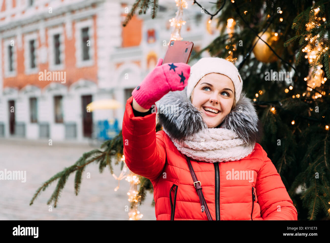 Riga, Lettonia. Giovane bella bella ragazza caucasica Donna vestita in rosso giacca e cappello bianco divertendosi e tenendo Selfie foto sullo smartphone sul retro Foto Stock