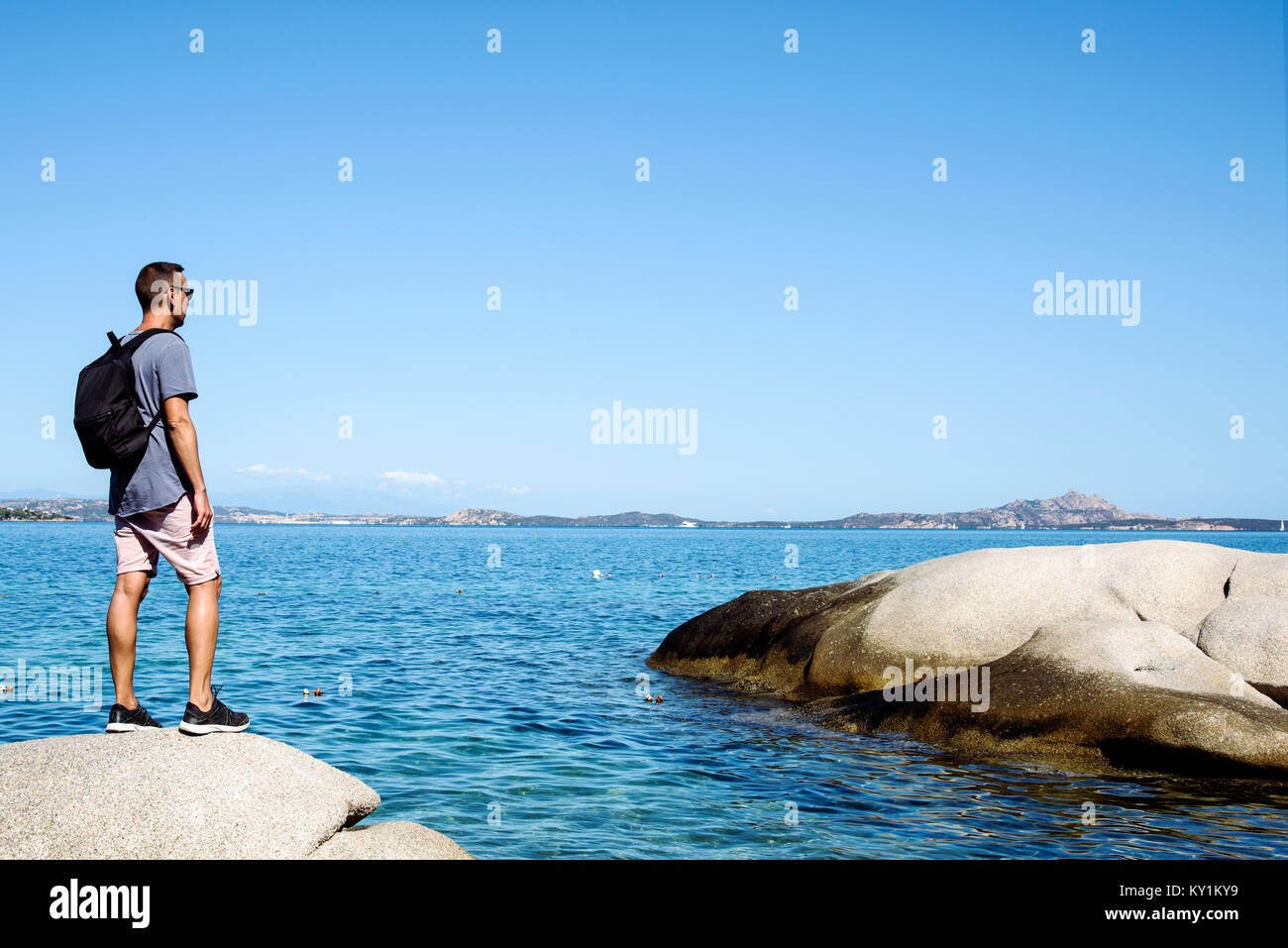 Un giovane uomo caucasico, visto da dietro, portando uno zaino osservando il mare in Costa Smeralda, in Sardegna, Italia, con un grande spazio vuoto nel Foto Stock