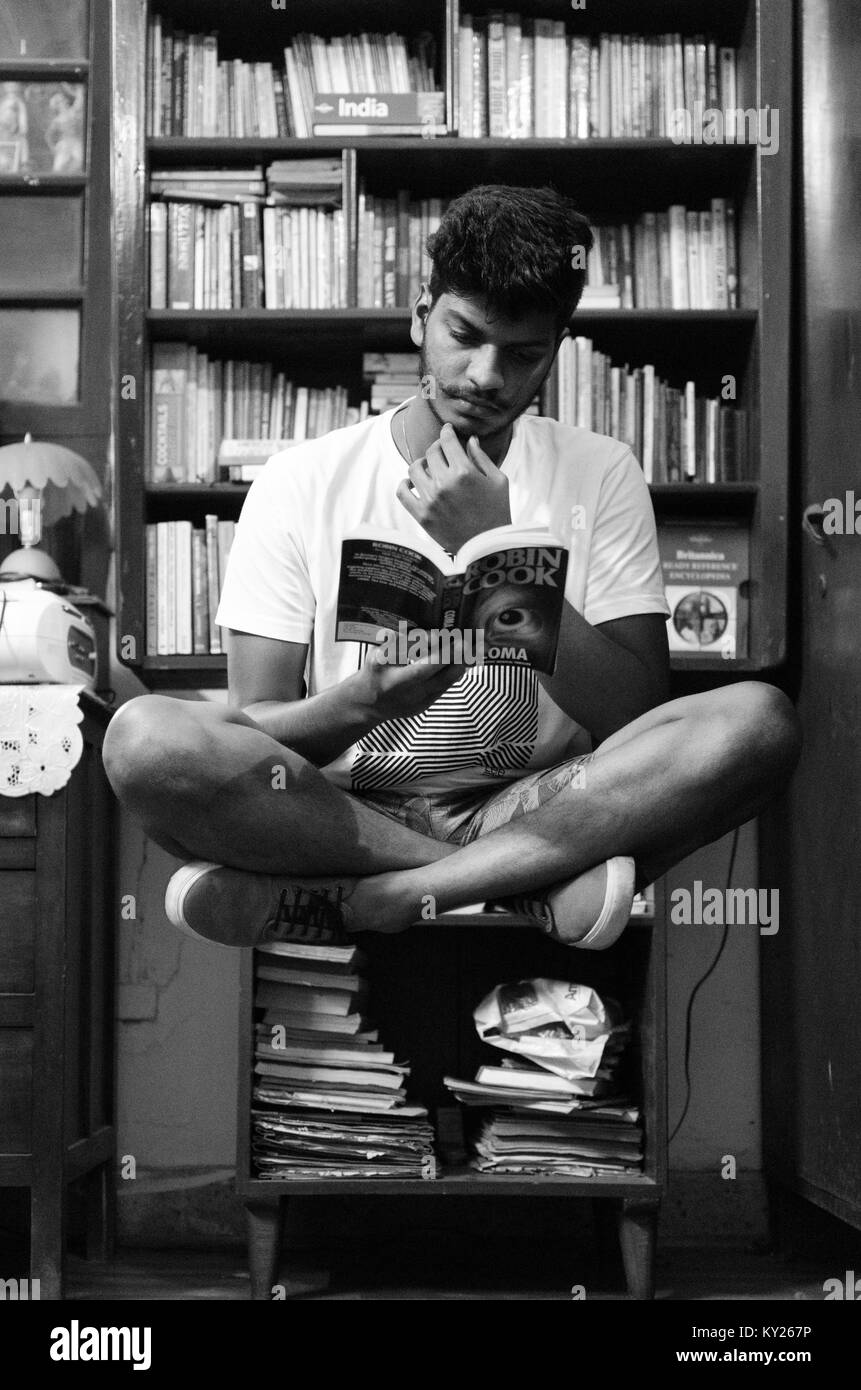 I giovani adulti ragazzo con le gambe incrociate, in posizione seduta, la levitazione davanti a uno scaffale, la lettura di un libro. Foto Stock