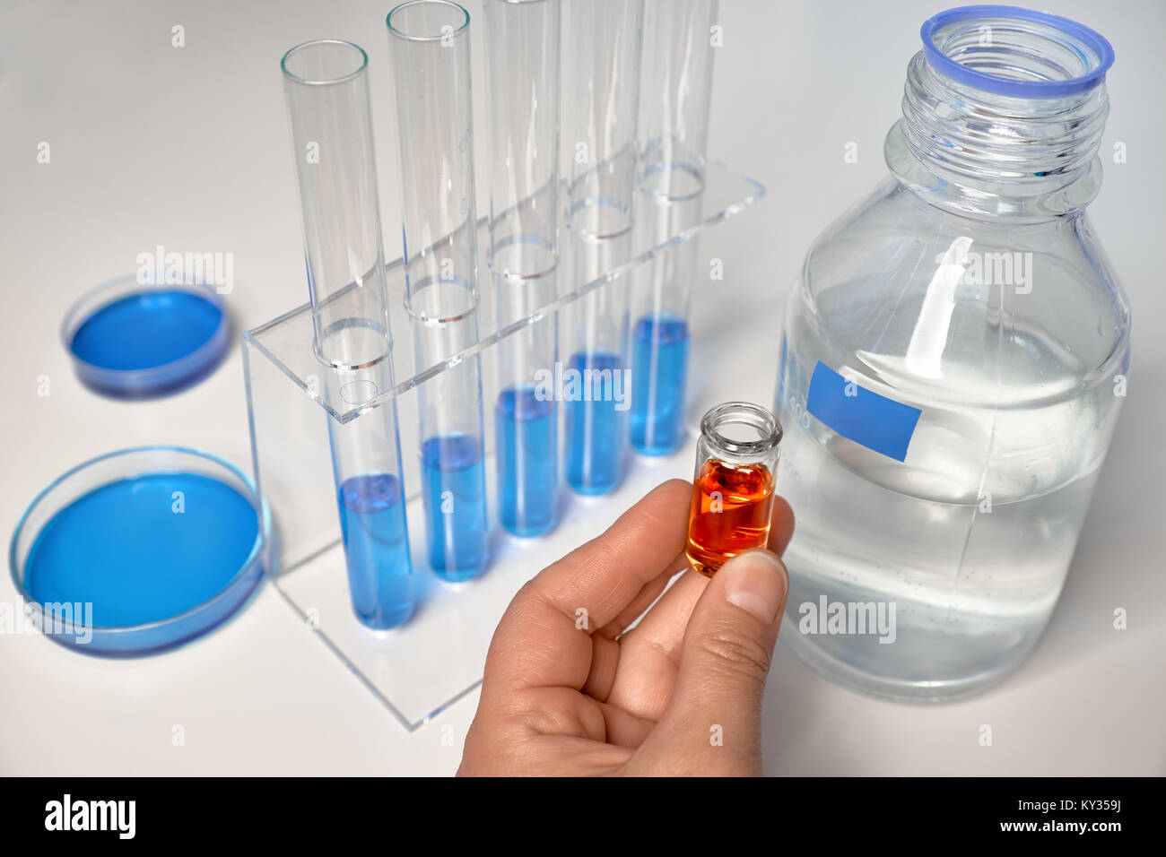 Background scientifico in blu, bianco e arancione. Il campione di liquido in mano femmina, blu campioni liquidi in tubi di vetro e piatti. Foto Stock