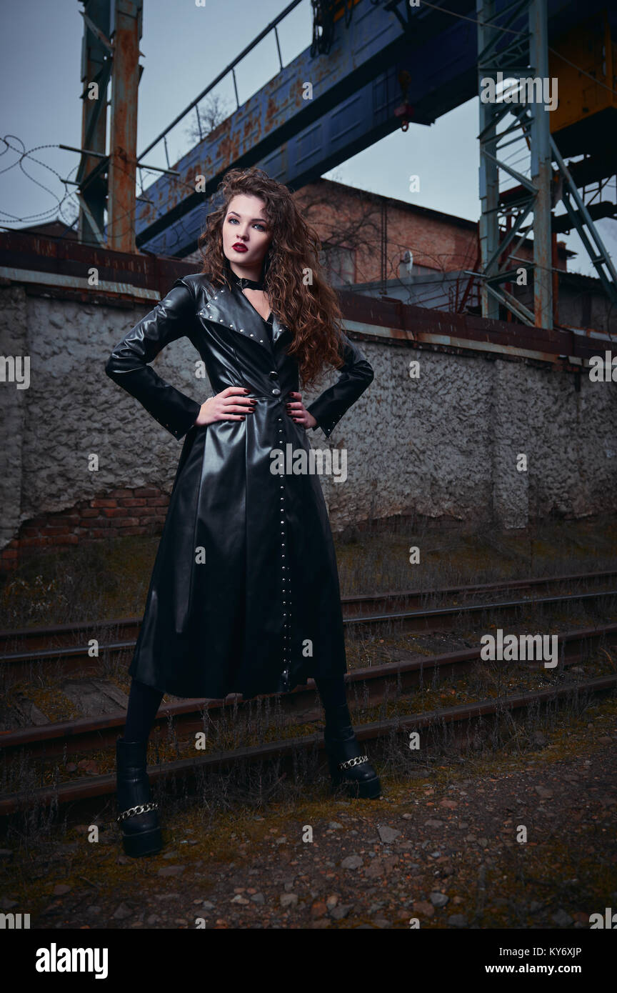 Moda shot: ritratto di una bella ragazza di goth (modello informale) in cappotto di pelle in piedi alla ferrovia (zona industriale) Foto Stock
