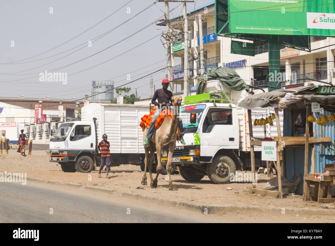 Un uomo che cavalca un cammello sul marciapiede lungo una strada, Nairobi, Kenya, Africa orientale Foto Stock