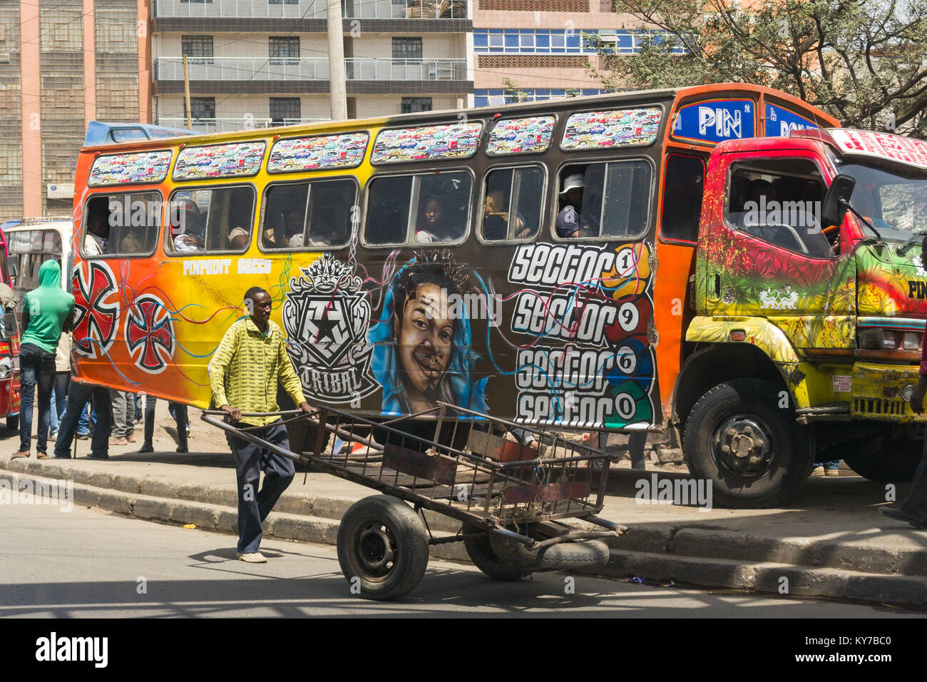 Un uomo spinge la sua mano carrello sulla strada oltre un decorate luminosamente bus si fermava sul marciapiede, Nairobi, Kenya, Africa orientale Foto Stock