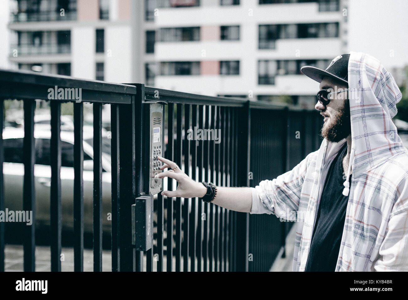 Giovane uomo con la barba in luce a scacchi in giacca con cappuccio in un cappello e occhiali da sole preleva l'appartamento numero sul tastierino per chiamare il citofono Foto Stock