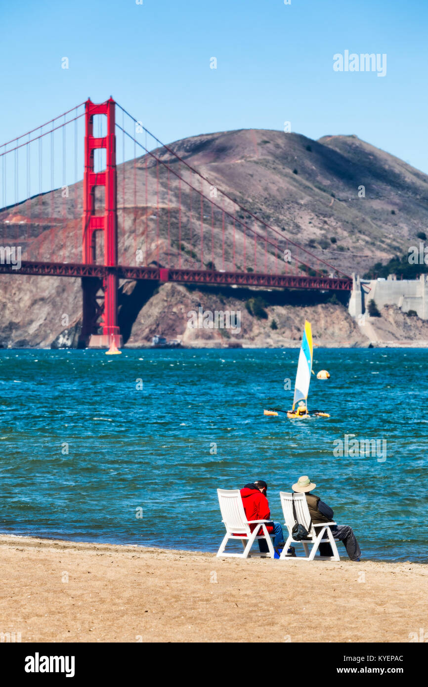 Due persone sedute a sdraio in spiaggia dall'acqua su una San Francisco Bay spiaggia sabbiosa. Golden Gate Bridge in background. Vista dei Promontori Marin. Foto Stock