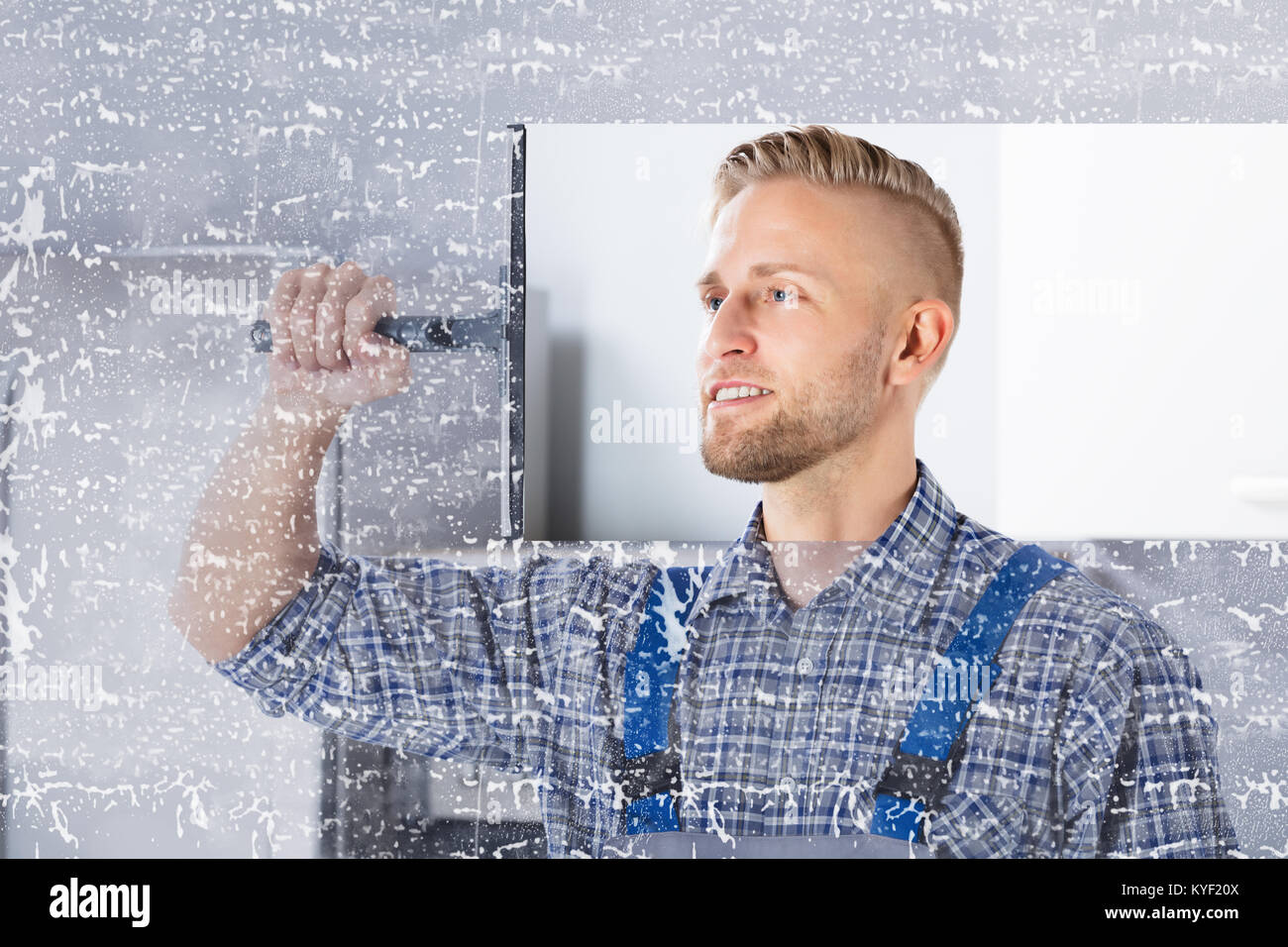 Sorridente giovane lavoratore Pulizia vetro Vetro con spatola in cucina Foto Stock