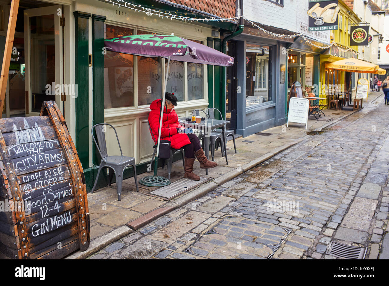 I vecchi tourist donna seduta al di fuori di un pub avente un caffè e la lettura in Mercery Lane, Canterbury Foto Stock