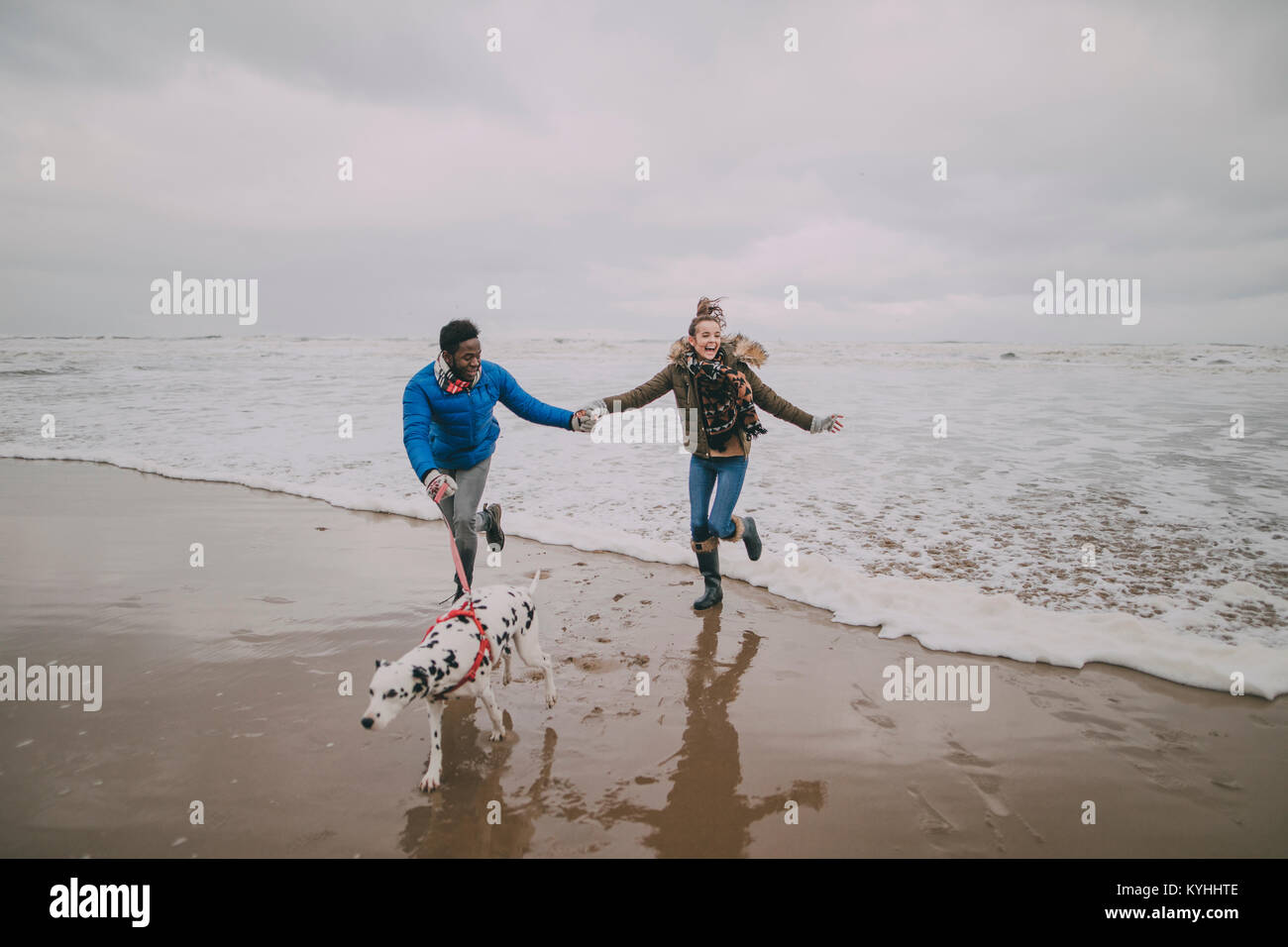 Una coppia giovane scappare dalle onde mentre si camminava il loro cane su una spiaggia invernale. Foto Stock