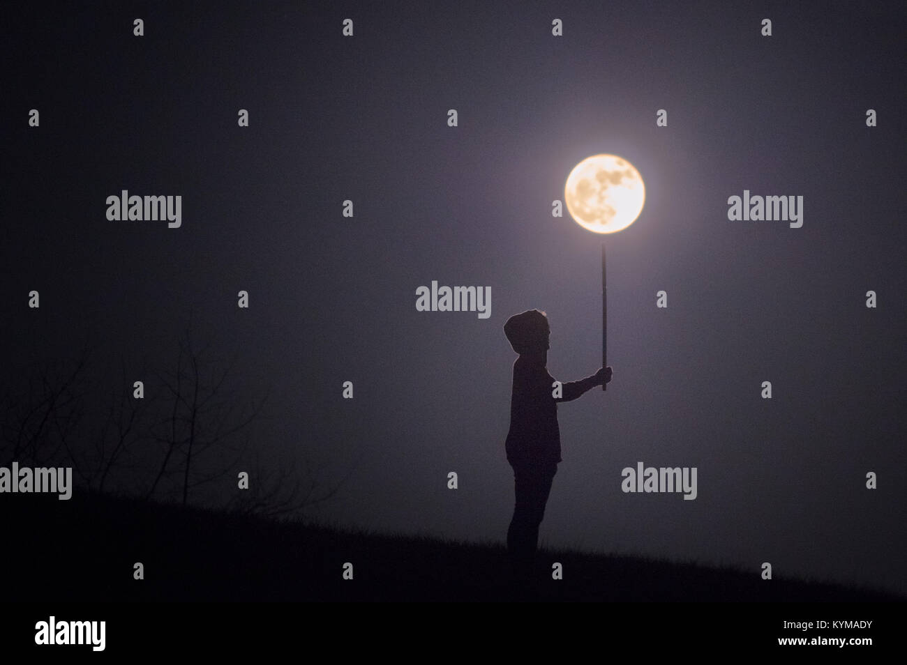 Una ragazza che indossa una felpa con cappuccio stagliano sorge circondato da tenebre sotto una luna piena che figurano per bilanciare la luna su un palo, come se tiene un gigante lig Foto Stock