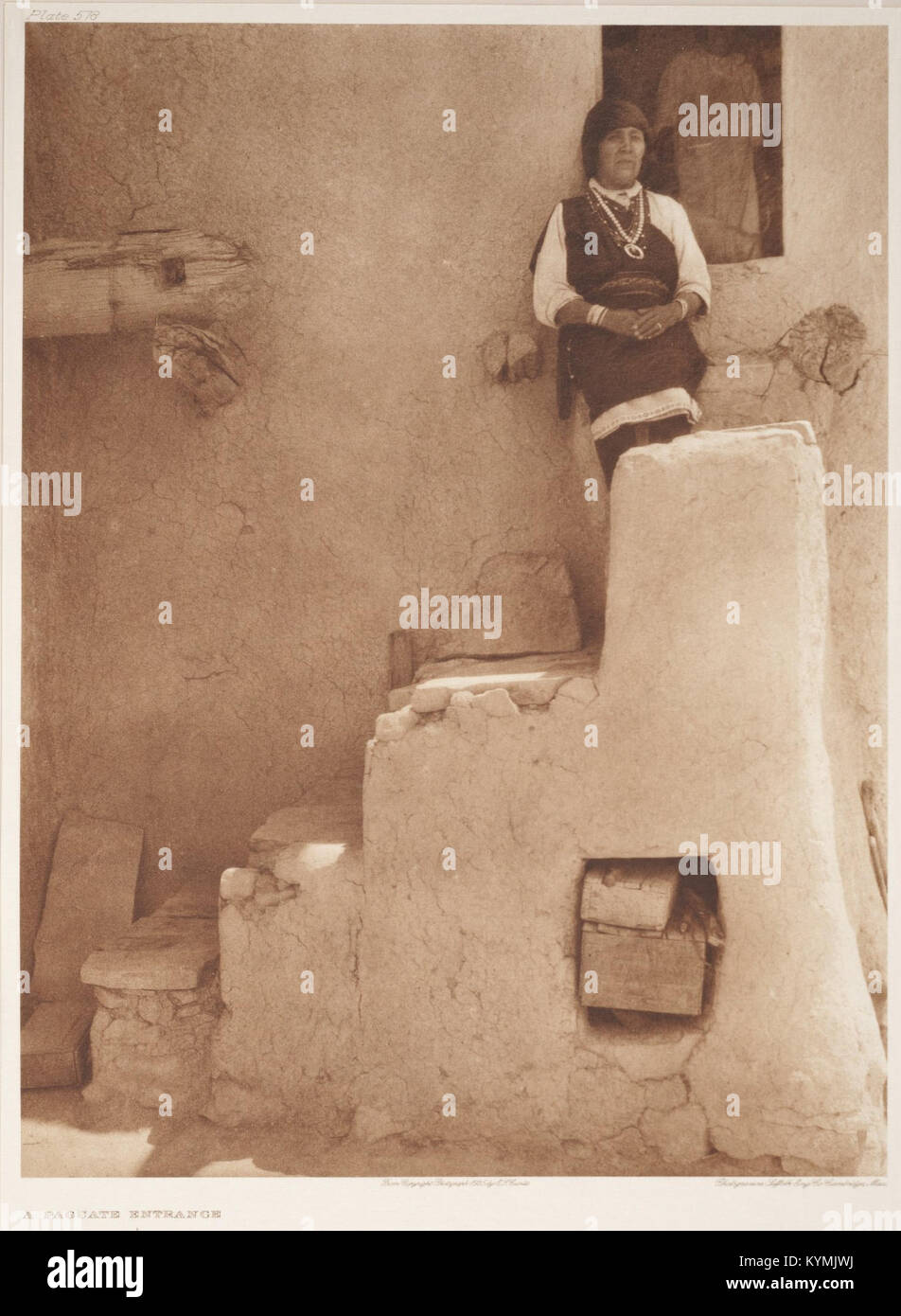Un'illustrazione storica di una donna nativa americana in abbigliamento tradizionale, in piedi di fronte a una casa di mattoni a Paguate, New Mexico. L'immagine fa parte della collezione Edward S. Curtis ospitata nelle Smithsonian Institution Libraries. La fotografia in bianco e nero mette in risalto i gioielli e i vestiti del soggetto in un'antica ambientazione del passato. Foto Stock