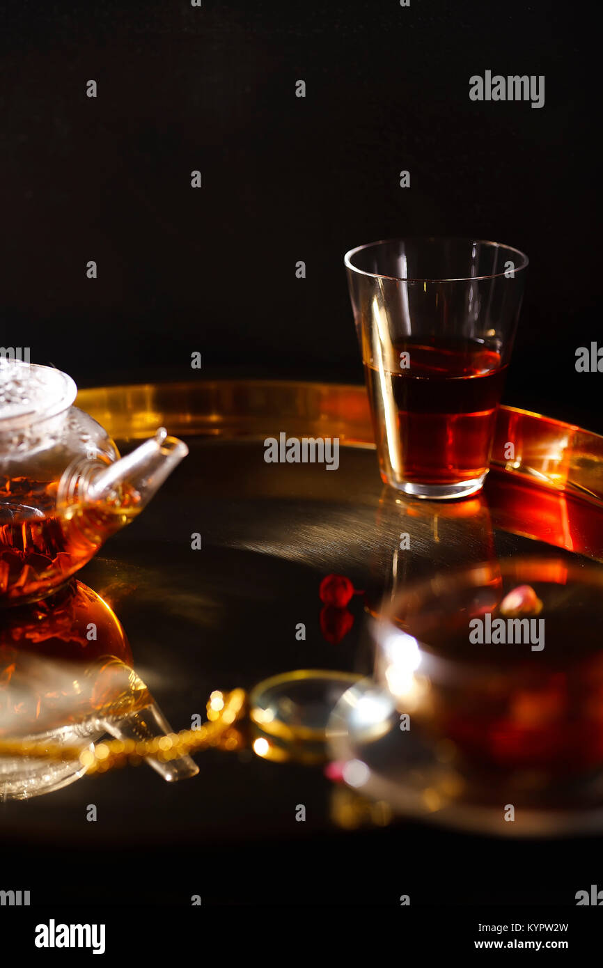 Piccola teiera di vetro e bicchieri con acqua calda tè nero, essiccati petali di rose, lente di ingrandimento tascabile sulla catena d'oro sul vassoio d'oro Luce della Sera. Foto Stock