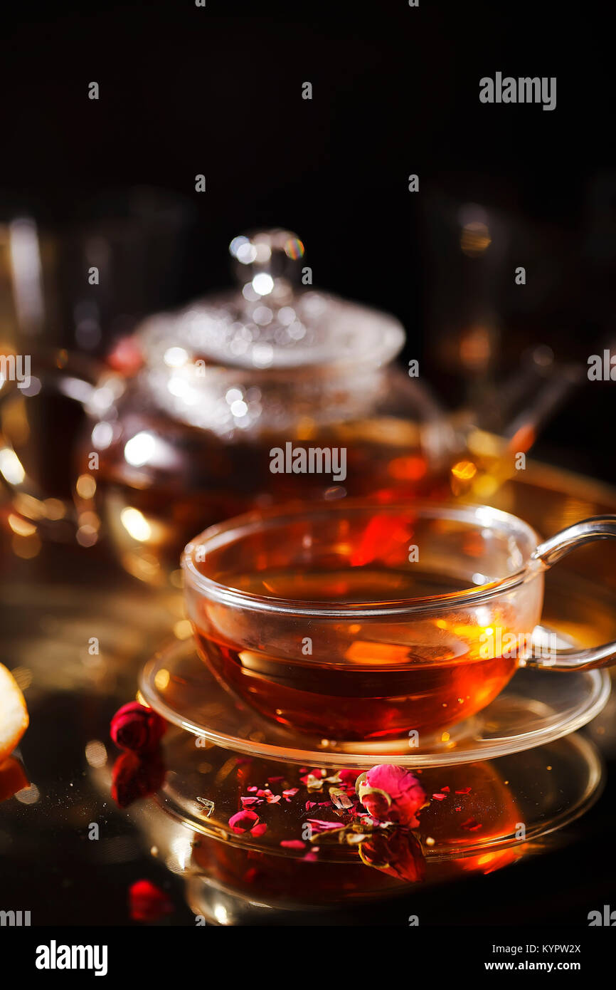 Verticale di close-up shot di piccolo bicchiere teiera e tazza calda con tè rosso, essiccati petali di rose, lente di ingrandimento tascabile sulla catena d'oro sul vassoio d'oro Sera lig Foto Stock