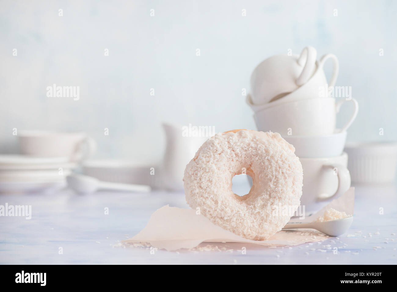 Ciambella con topping di cocco su uno sfondo luminoso con cottura carta oleata e tazze in porcellana e piattini. Elevato La chiave fotografia di cibo. Pila di tazze. Foto Stock