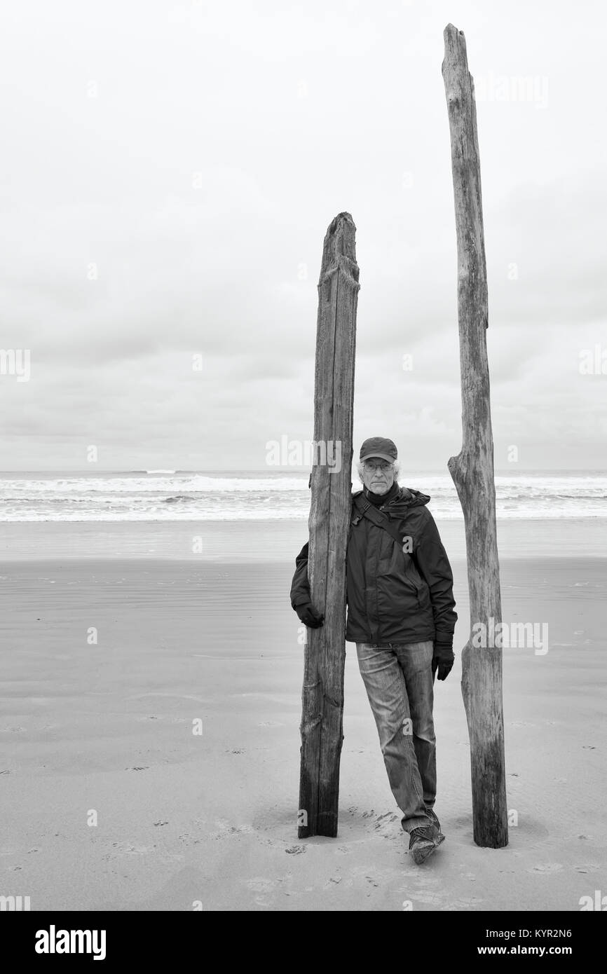 Ritratto di senior uomo sulla spiaggia in piedi con driftwood posti nella sabbia. Ocean in background. Bianco e nero monocromatico. Copia dello spazio. Foto Stock