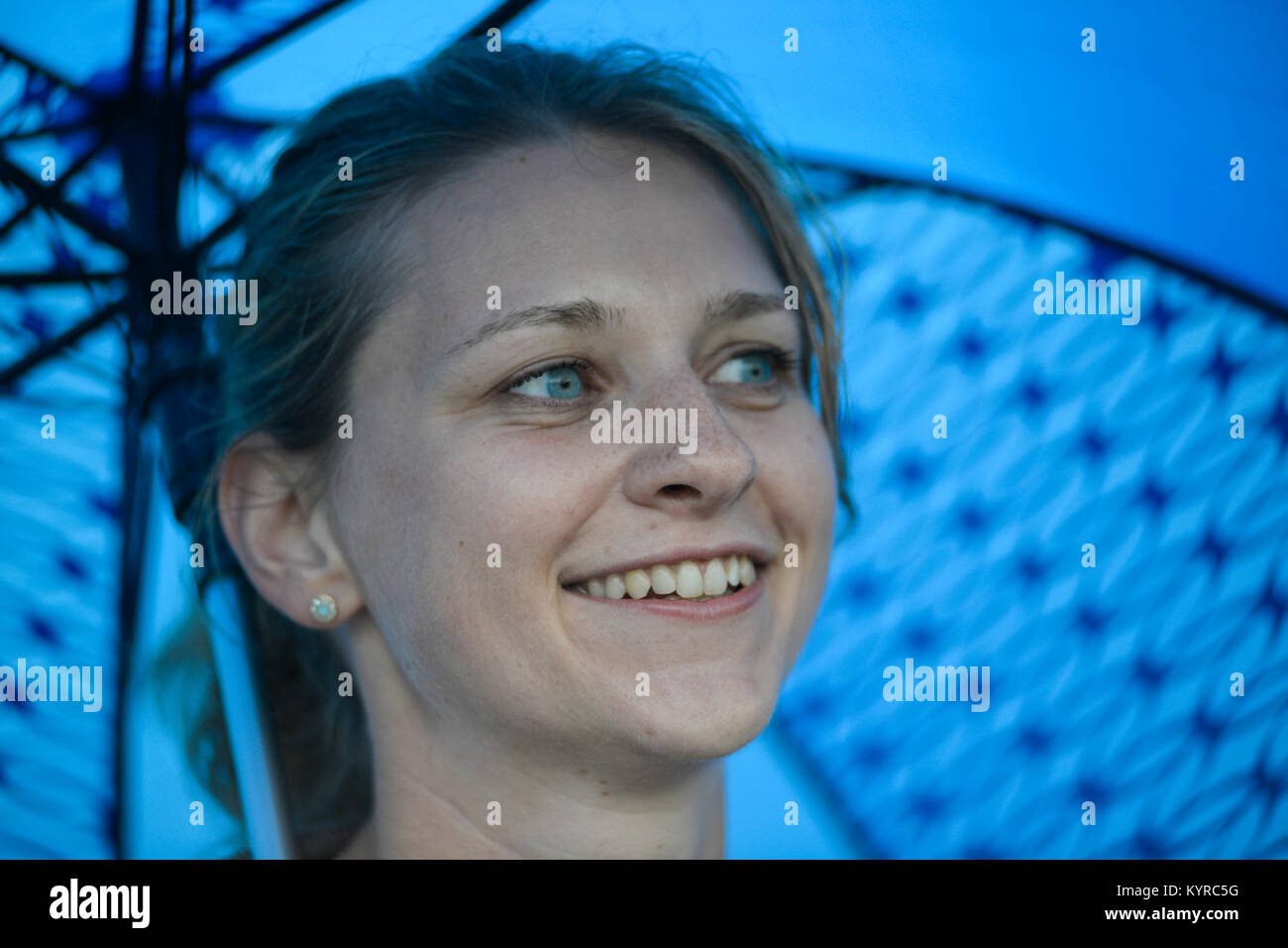 Una ragazza bionda con gli occhi blu sorridente sotto un ombrello blu Foto Stock