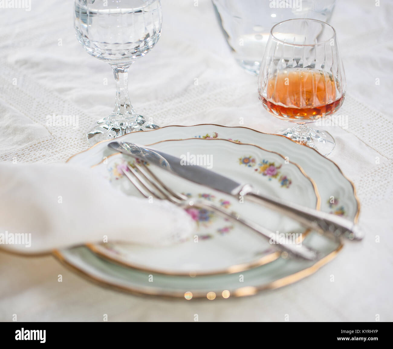 Lusso piatti di porcellana, argenteria giacendo attraverso i piatti e bicchieri di cristallo e un bicchiere di cognac su bianco vintage tovaglia di pizzo Foto Stock