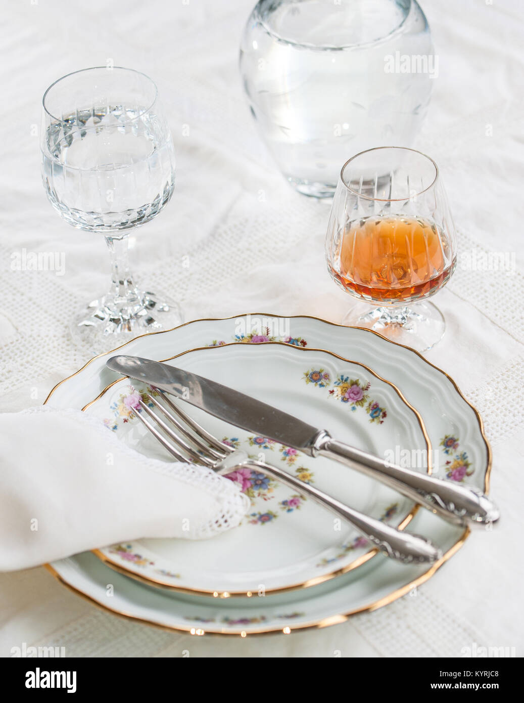 Lusso piatti di porcellana, argenteria giacendo attraverso i piatti e bicchieri di cristallo e un bicchiere di cognac su bianco vintage tovaglia di pizzo Foto Stock
