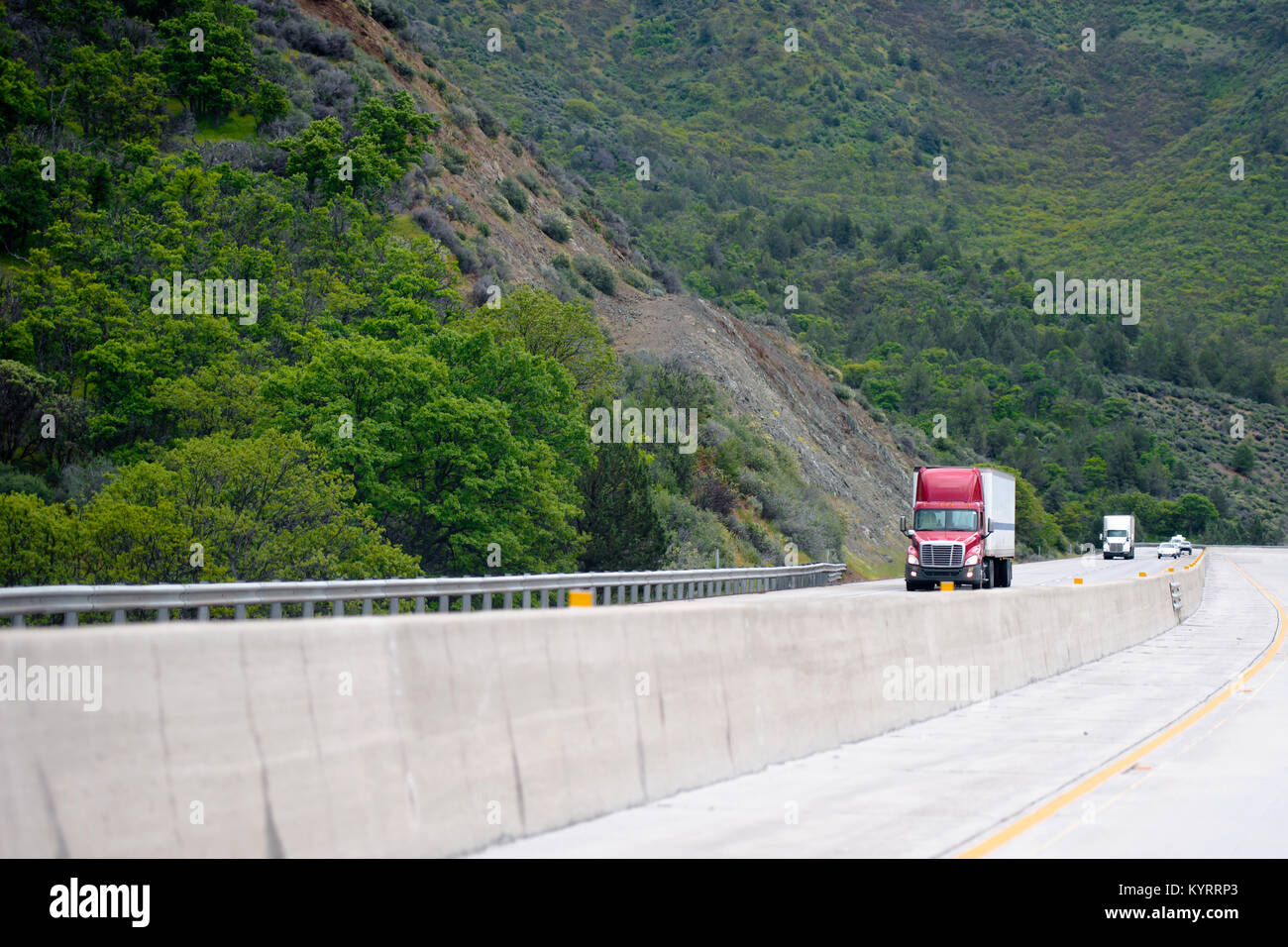 Red big rig semi camion con rimorchio andando su per la collina su avvolgimento in mountain pass autostrada con un altro camion e automobili traffico e foresta verde Foto Stock