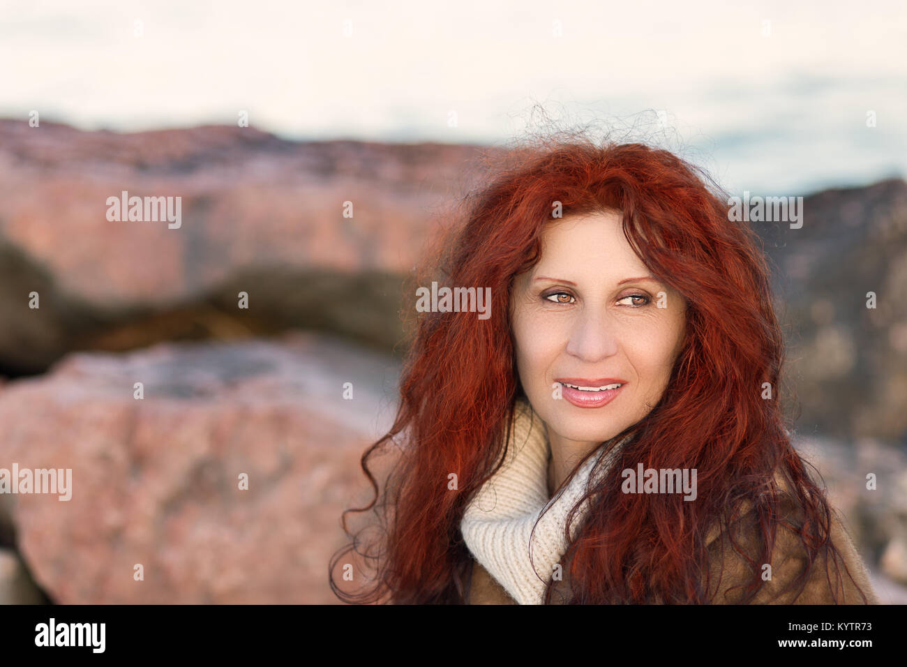 Affascinante di mezza età donna sorridente seduto sulle rocce con onde in background in Italia in una fredda giornata invernale Foto Stock