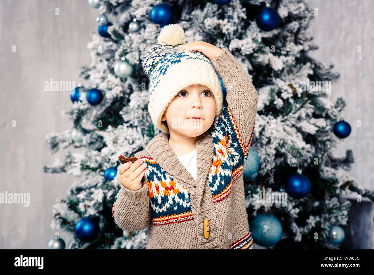 Piccolo ragazzo divertente nel copricapo e corpo maglione di lana mangia i  dolci al cioccolato sullo sfondo di un albero di Natale alla vigilia di  Natale home.mangiò e cosparse le sue guance,