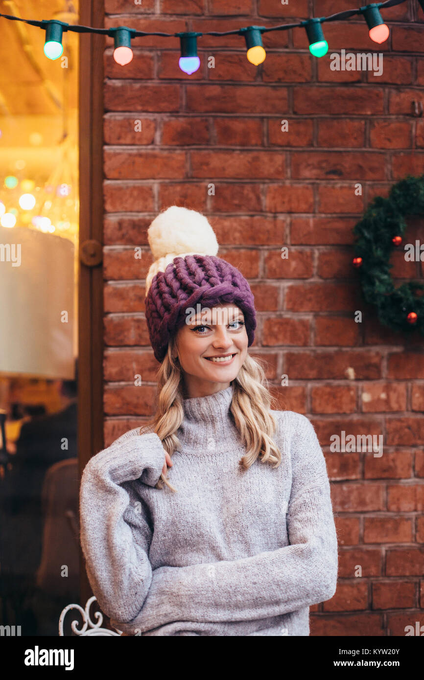 Donna bionda in maglia viola cappelli e maglione grigio rendono posa con  muro di mattoni e una ghirlanda di Natale sullo sfondo Foto stock - Alamy