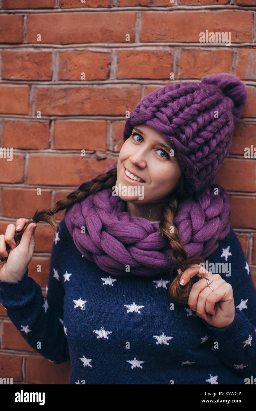 Brunette donna in viola berretto lavorato a maglia, lavorata a maglia  Sciarpa infinity e maglione blu con stelle bianche su di esso fare in posa  con un muro di mattoni sullo sfondo