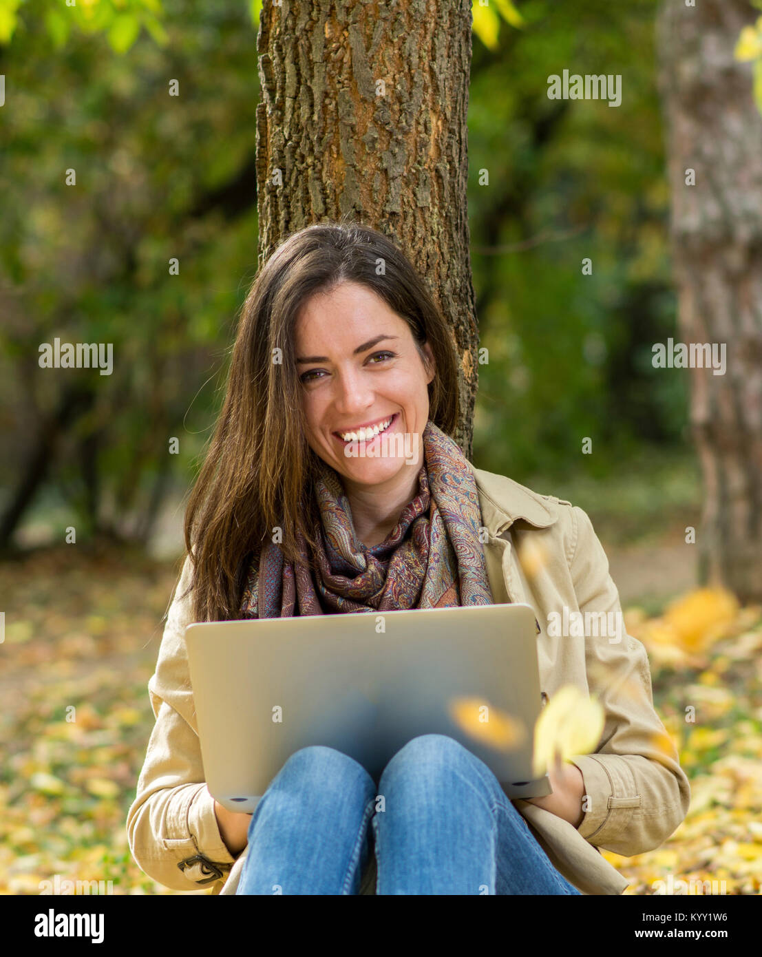 Ritratto di donna sorridente utilizzando il computer portatile durante la seduta di tree durante l'autunno Foto Stock