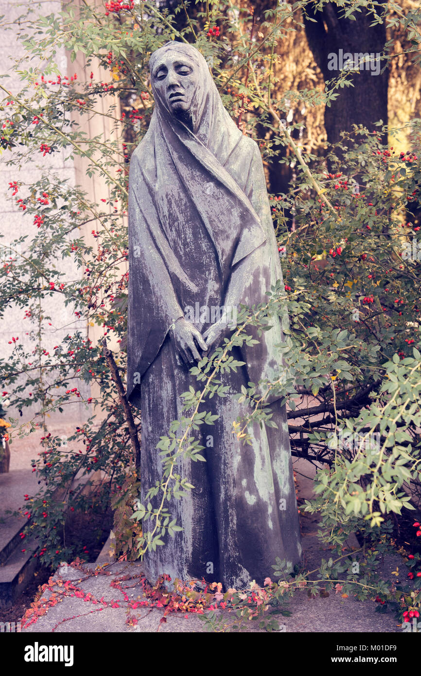 Una statua solenne, intemprata, di una figura occultata in un giardino, circondata da rami sovracresciuti e bacche rosse, che evocano temi di malinconia, Foto Stock Una statua solenne, intemprata, di una figura occultata in un giardino, circondata da rami sovracresciuti e bacche rosse, che evocano temi di malinconia, Foto Stock