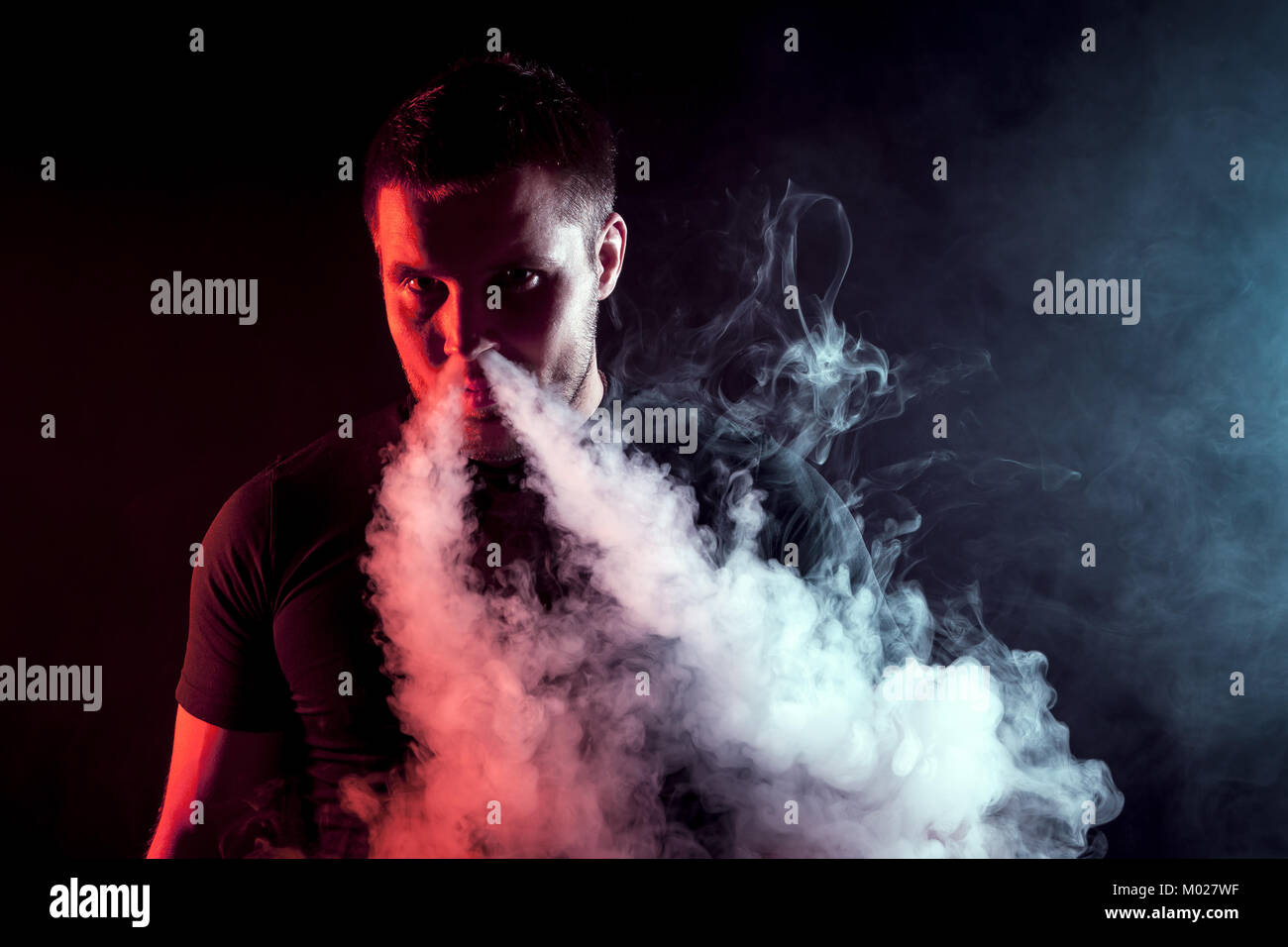 Giovane maschio elegante smoker in black T-shirt tirate fuori il suo naso e una grande nuvola di fumo da vape su un nero sfondo isolato Foto Stock