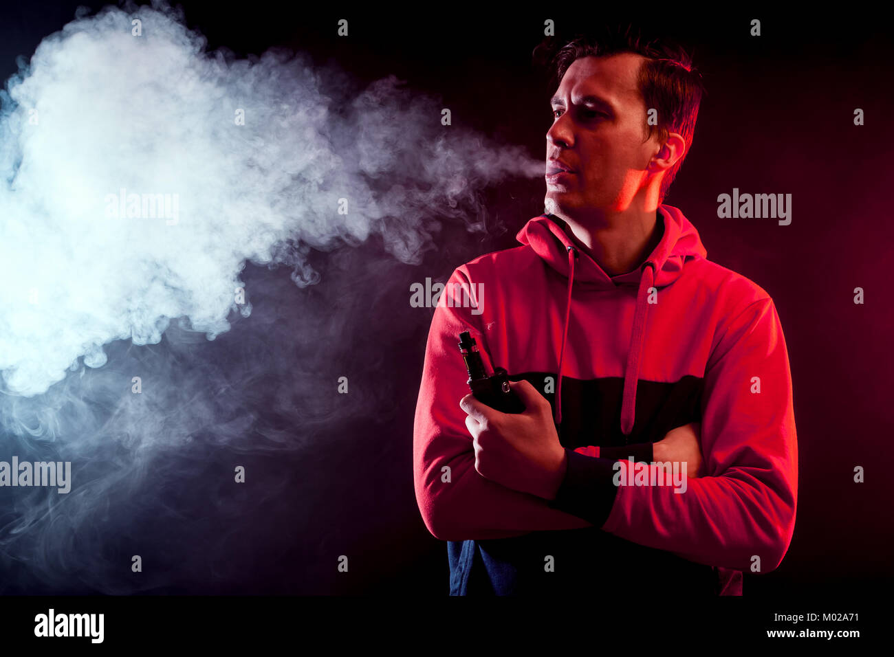 Giovane maschio elegante smoker in una felpa si brucia grande nube di fumo da vape al buio su un sfondo isolato Foto Stock