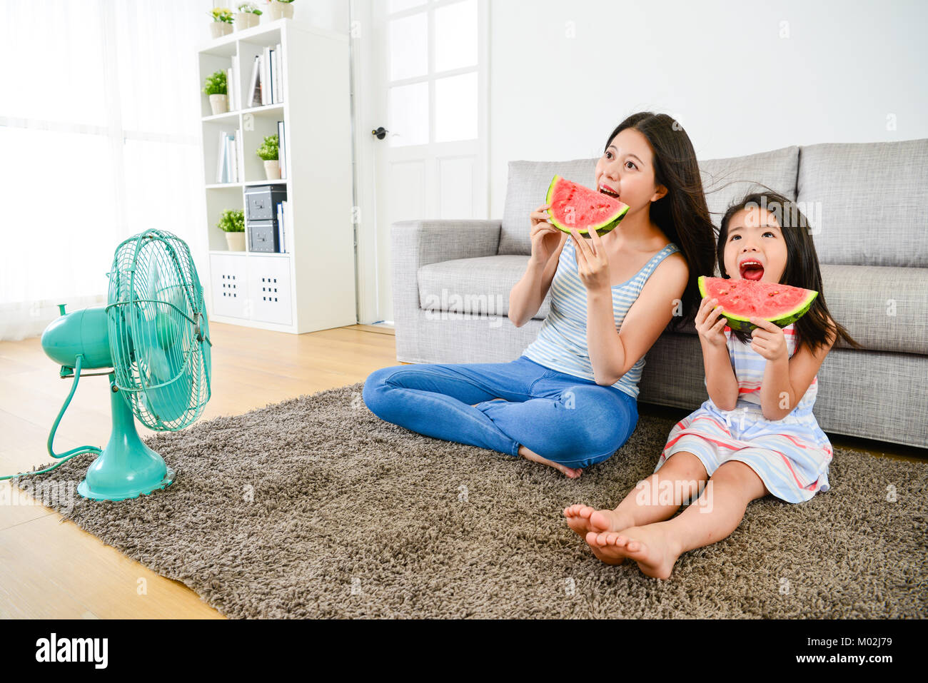 Bella Donna sorridente con giovani carino bambina mangiando anguria insieme in estate e soffiare la ventola elettrica rinfrescante. Foto Stock