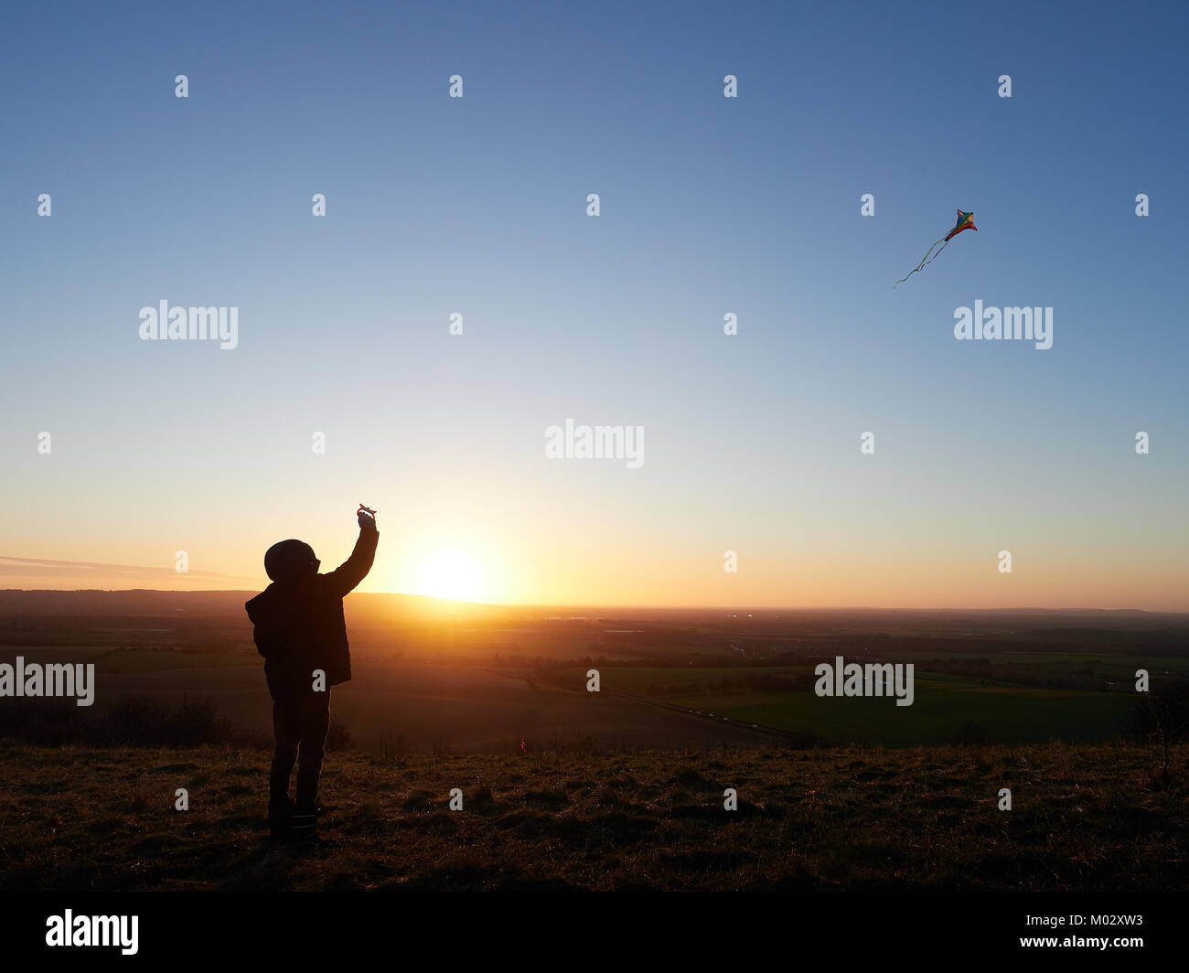 Bambino volare un aquilone in azzurro del cielo sopra la campagna Britannica stagliano contro il sole di setting oltre l'orizzonte Foto Stock