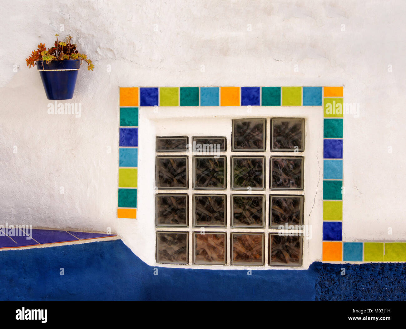 Piastrelle colorate intorno a una finestra di mattoni in vetro impostato nel muro bianco con un blu plantpot Foto Stock