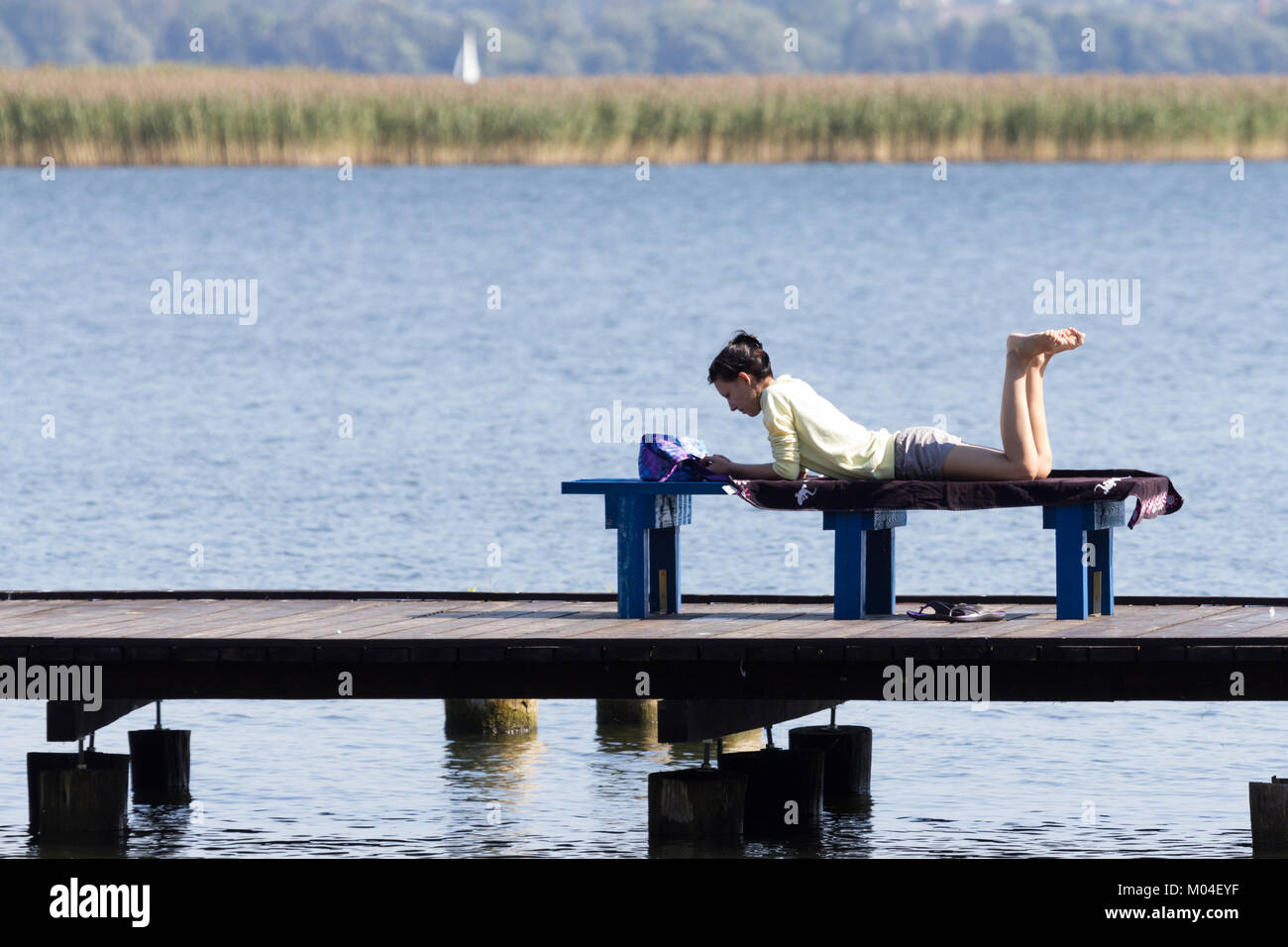 La donna è sdraiato su una panchina in riva al lago e la lettura di un libro. Wilkasy, la Masuria, grande zona dei laghi, Polonia Foto Stock