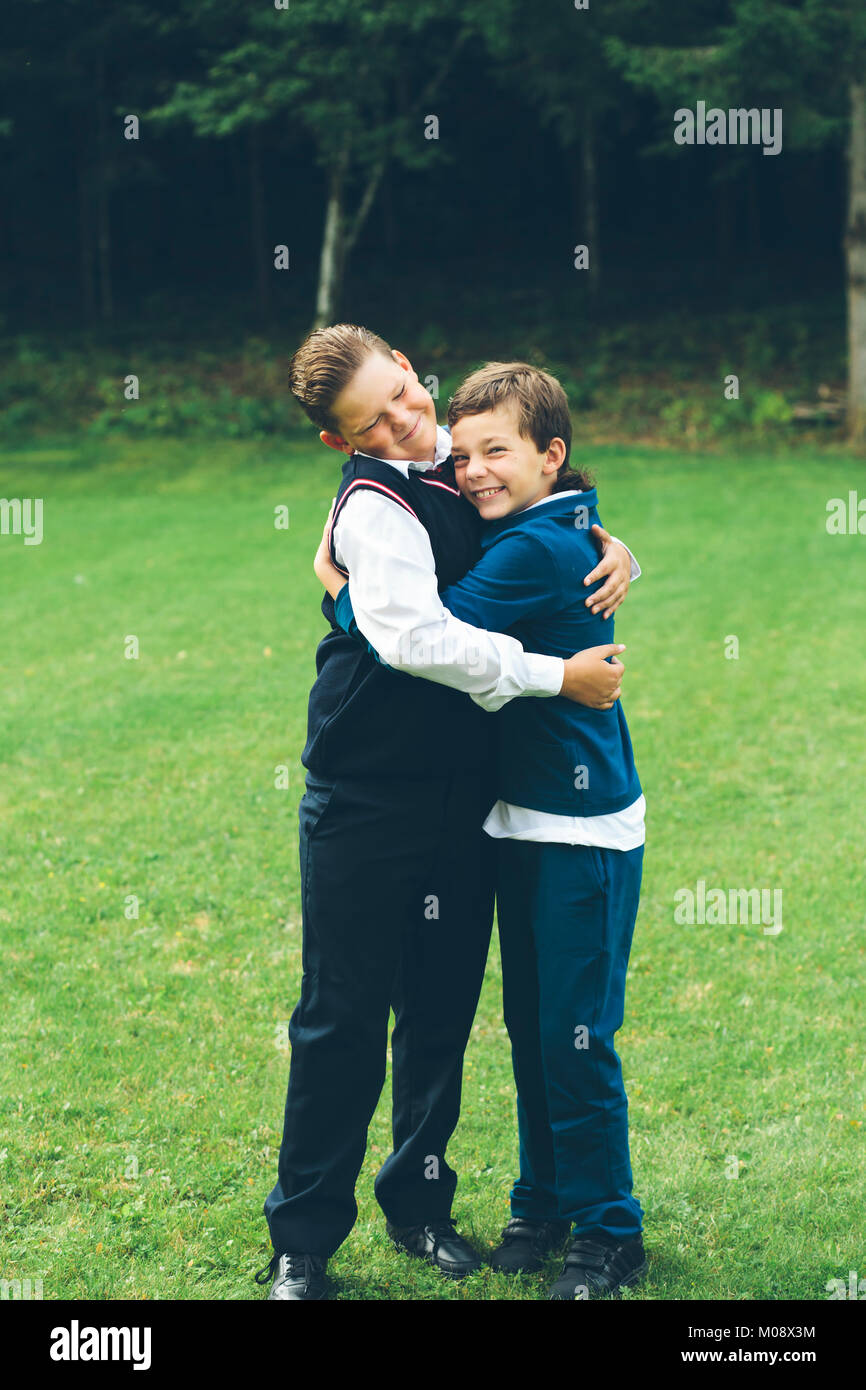 Due ragazzi, fratelli, scolari vestito in convenzionale che abbraccia ogni altro di fronte a una foresta in un prato in estate. Foto Stock