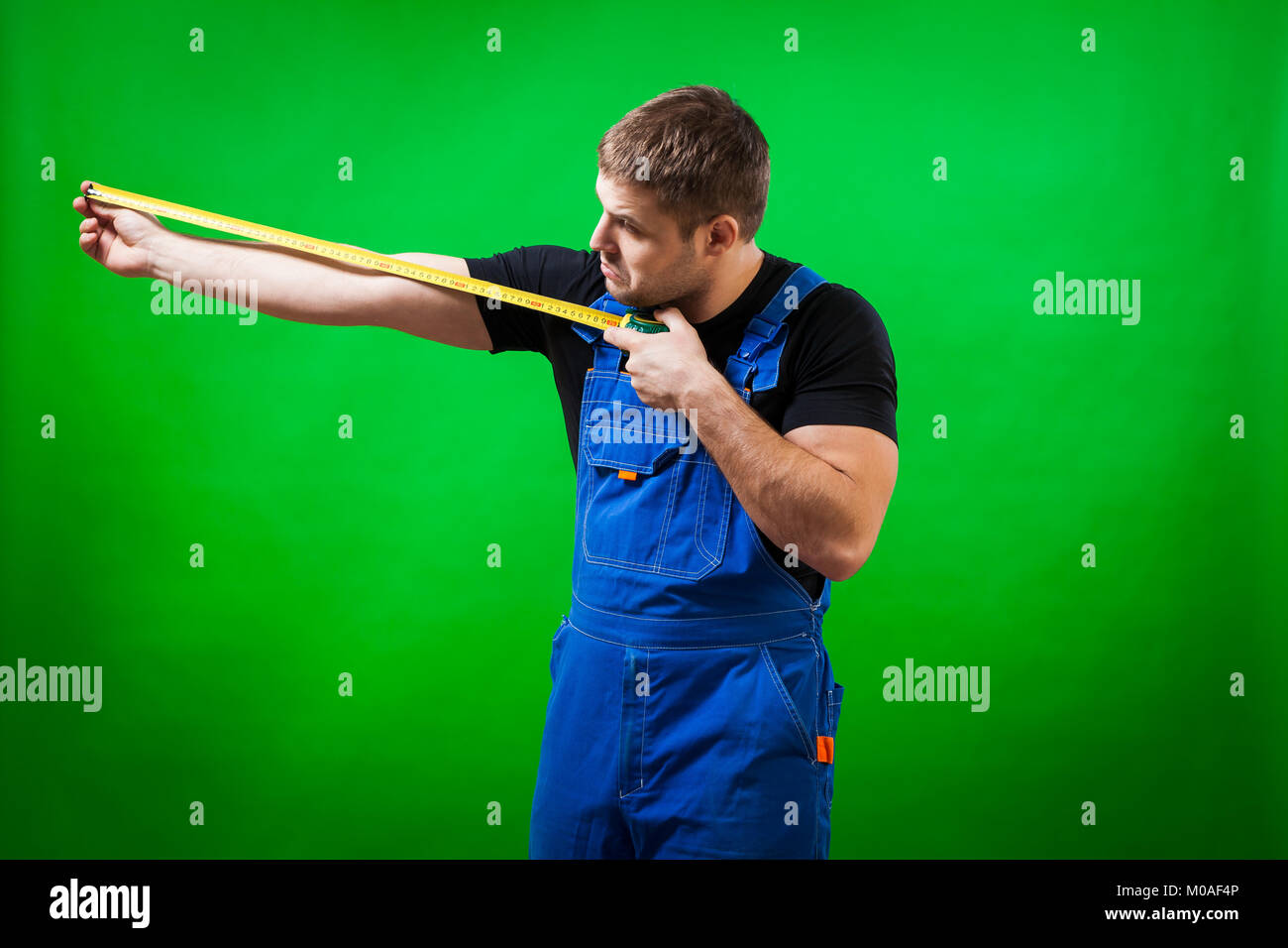 Un dai capelli scuri builder maschio in una T-shirt nera e blu tuta costruzione dimostra e misure con il suo lungo nastro giallo regola la sua mano su di una gr Foto Stock