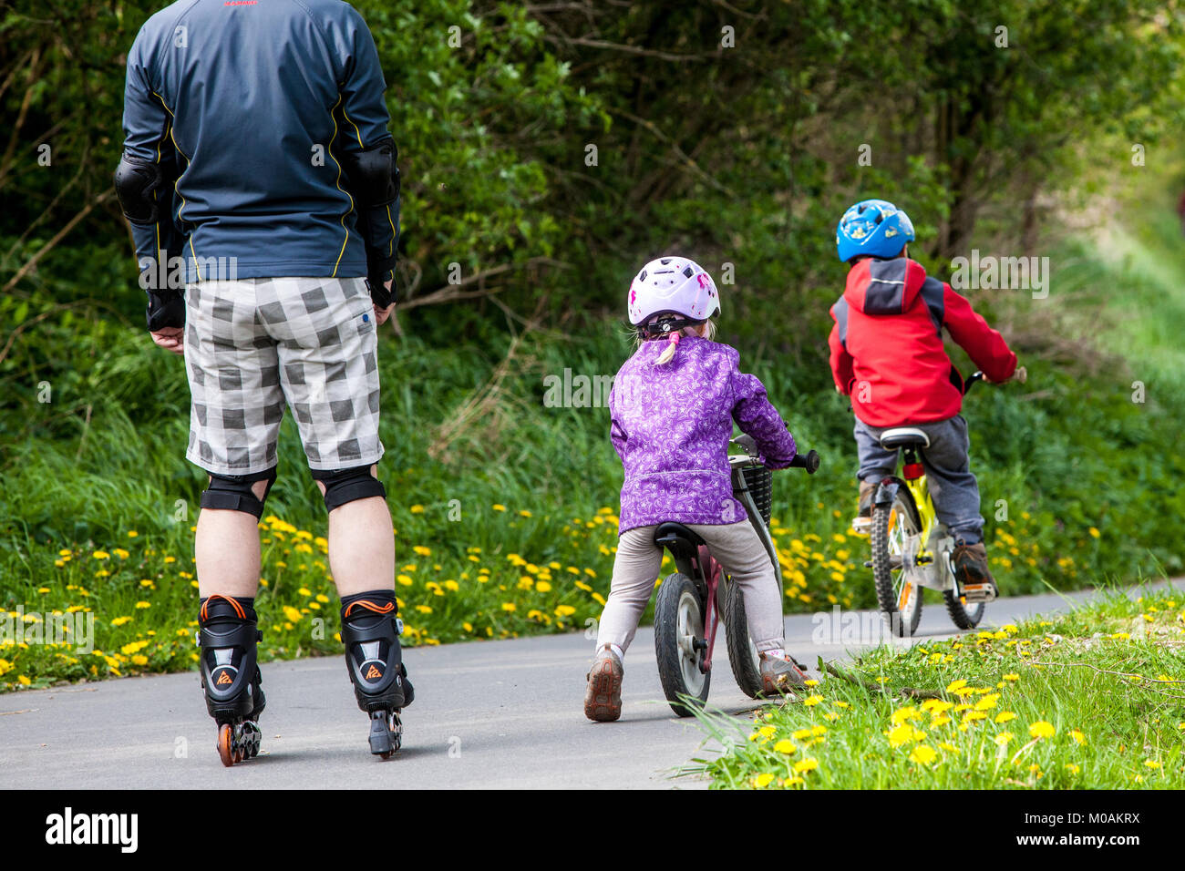 Bambini sotto la supervisione dei genitori due bambini vanno in bicicletta e padre su pattini a rotelle Foto Stock
