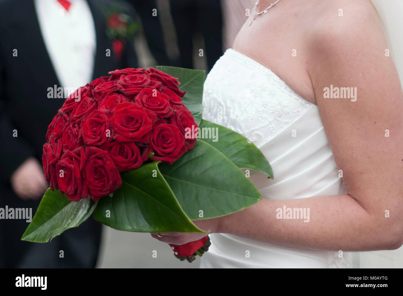 Sposa in abito bianco azienda rosso bouquet di fiori con lo sposo in background Foto Stock
