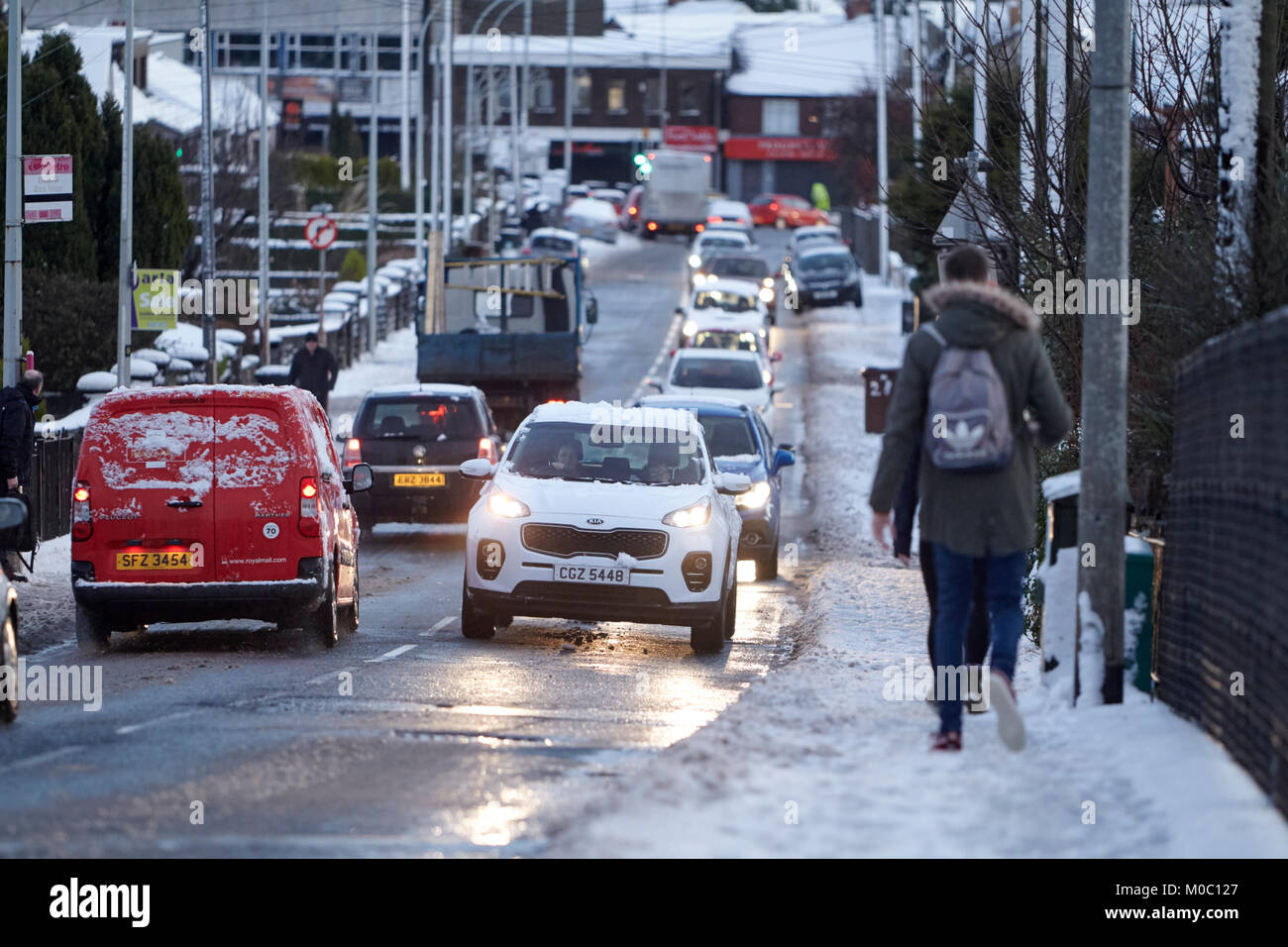 Il traffico mattutino con vetture di guida e la gente che cammina lungo la strada coperta di neve a Newtownabbey Irlanda del Nord Foto Stock