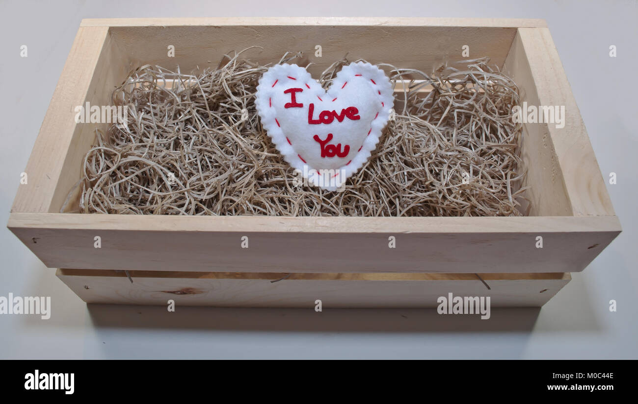 Un cuore bianco con "ti amo", scritto in tutta la parte anteriore di esso posa in un letto di paglia in una scatola di legno con uno sfondo bianco Foto Stock