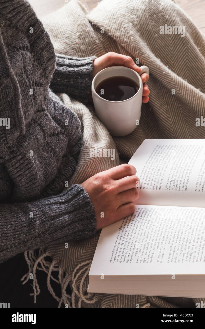 Donna che indossa lanosi grigio cardigan gustando una tazza di tè e la lettura di un libro sotto una coperta accogliente Foto Stock