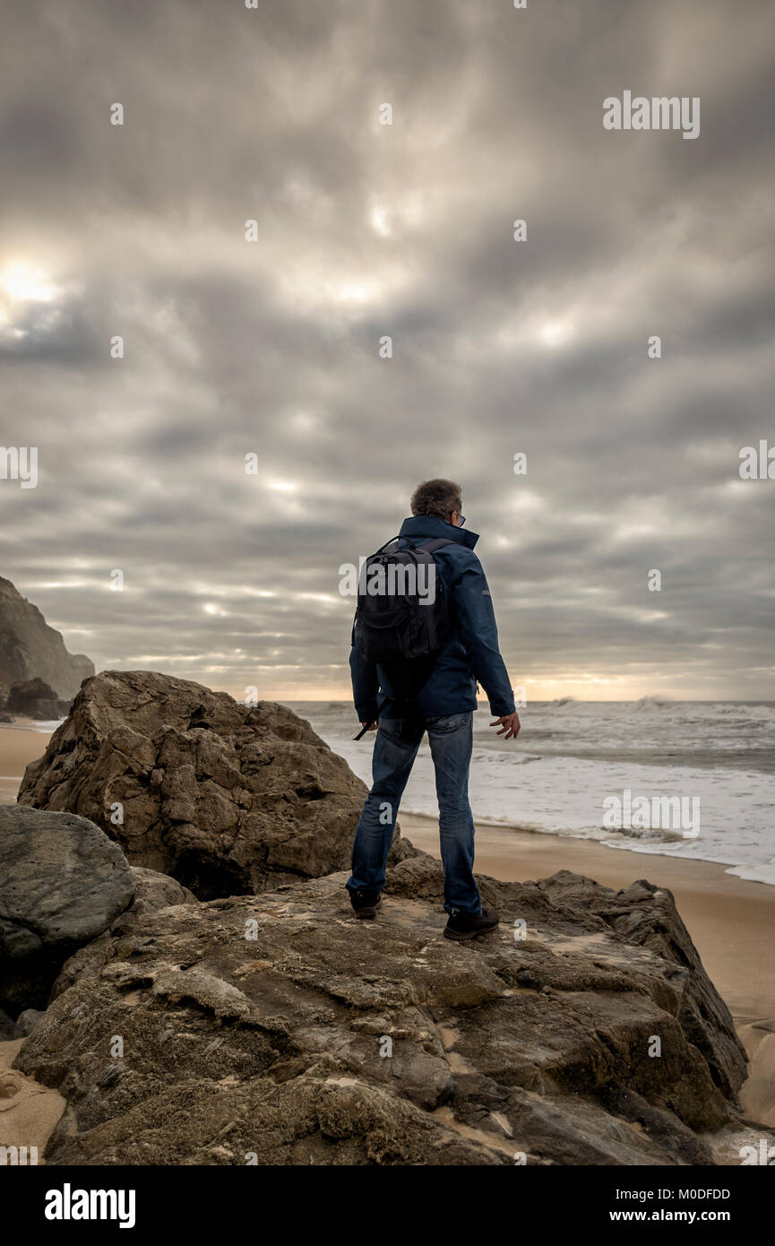 Uomo che indossa uno zaino in piedi sulle rocce affacciato sull'oceano. Foto Stock