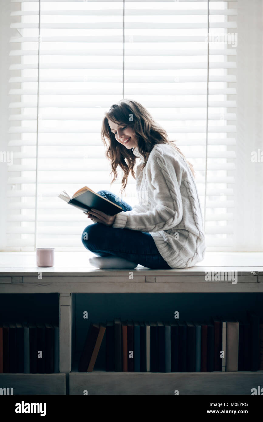 Felice donna bella lettura libro seduti sul davanzale di casa. Concetto di relax Foto Stock