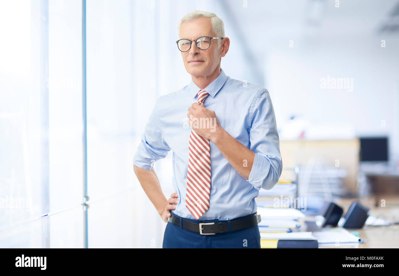 Imprenditore Senior indossando arrotolato shirt maniche e regolando la sua cravatta contro mentre in piedi presso l'ufficio. Foto Stock