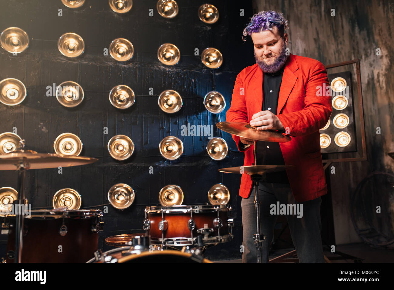 Batterista barbuto in tuta rossa sul palco con luci, in stile retrò. Esecutore musicale con i capelli colorati Foto Stock