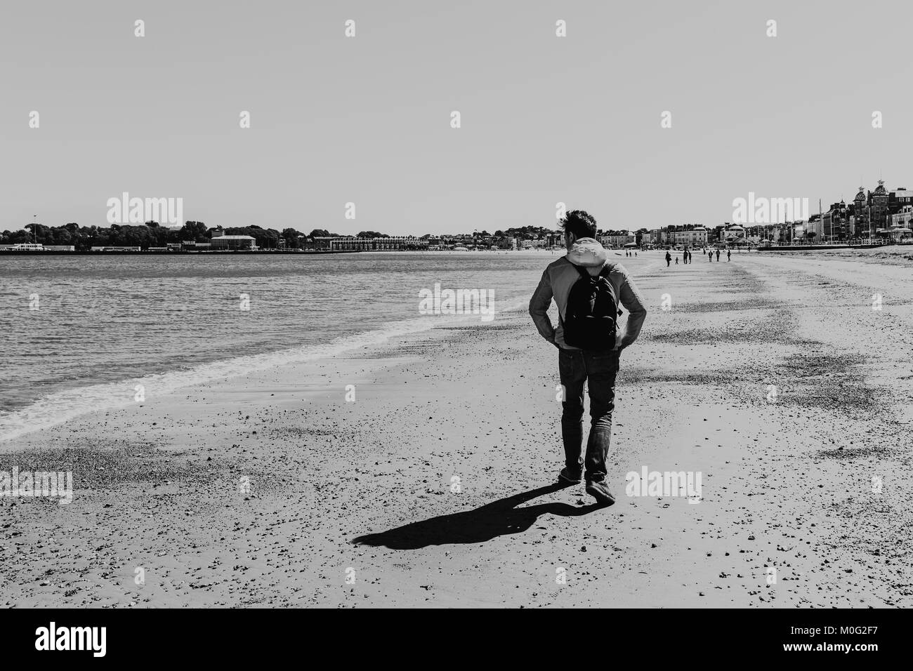 Giovane Uomo in camicia rossa e jeans camminando sulla spiaggia di Weymouth Dorset, Inghilterra in una limpida giornata di sole. Foto Stock