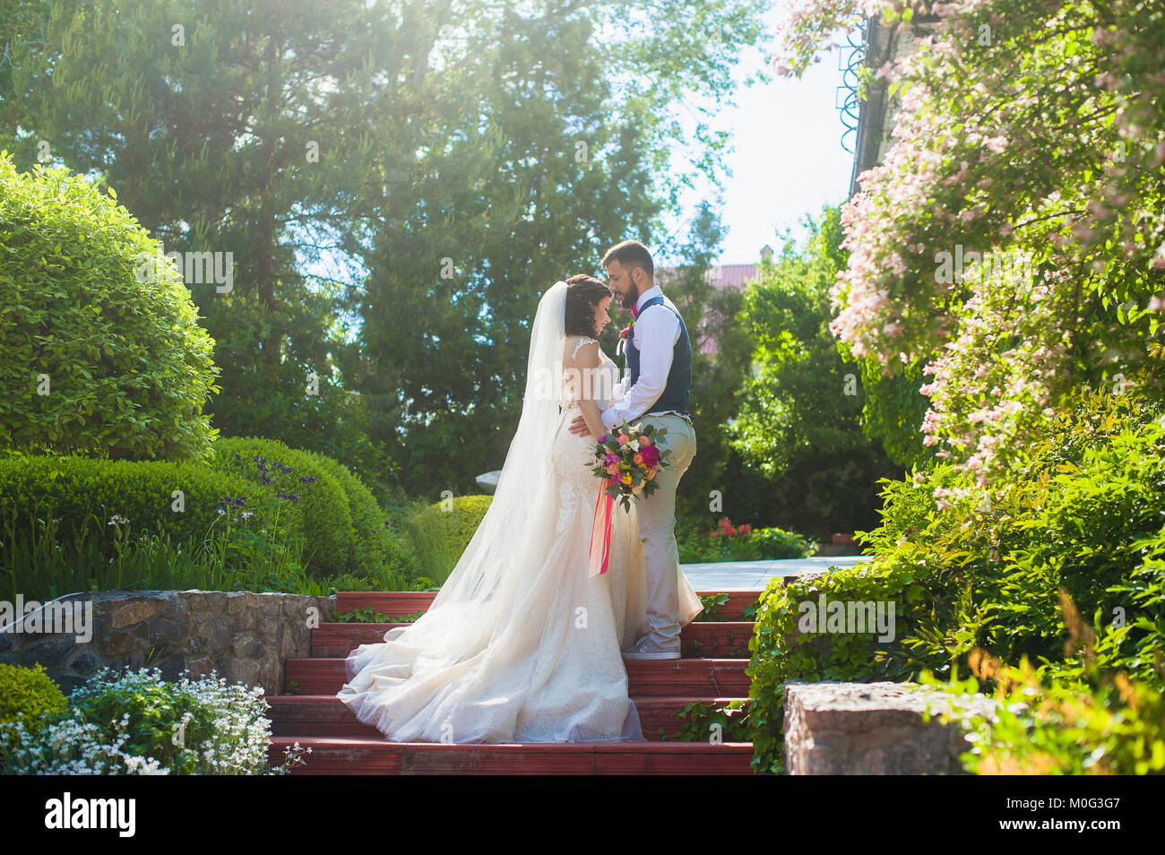 Bellissima proprio felice coppia sposata baciare. Sposa in bianco tromba  Abito in pizzo e lo sposo in giubbotto blu e sneakers in piedi in estate  verde parco sul th Foto stock -